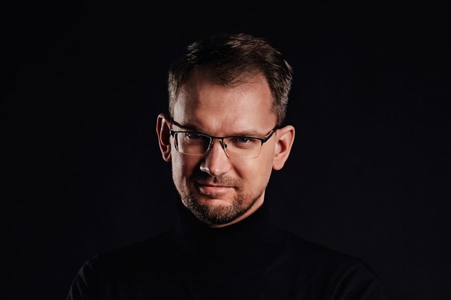 Marcin Dobrzyński in a black turtleneck, arms crossed, against a dark background